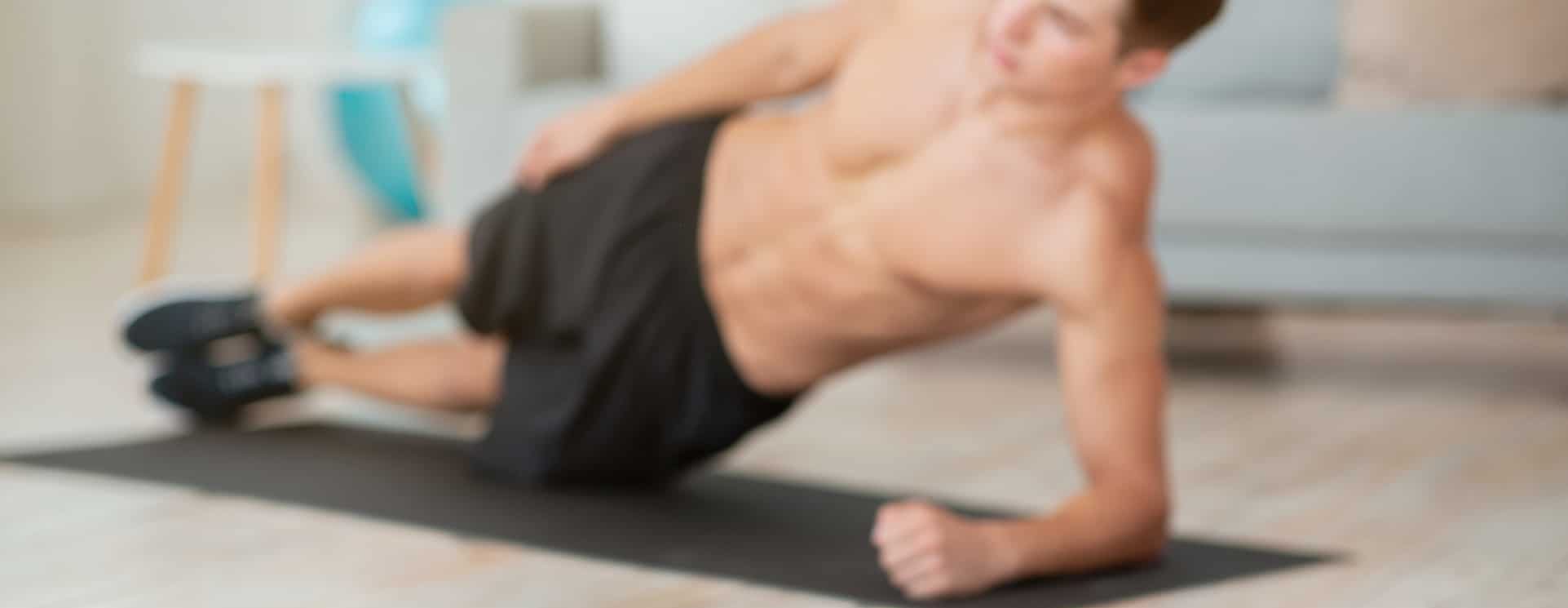 Dumbbells And Glass For Protein Shake Close Up, Guy Shirtless Makes Side Plank, On Mat In Living Room Interior